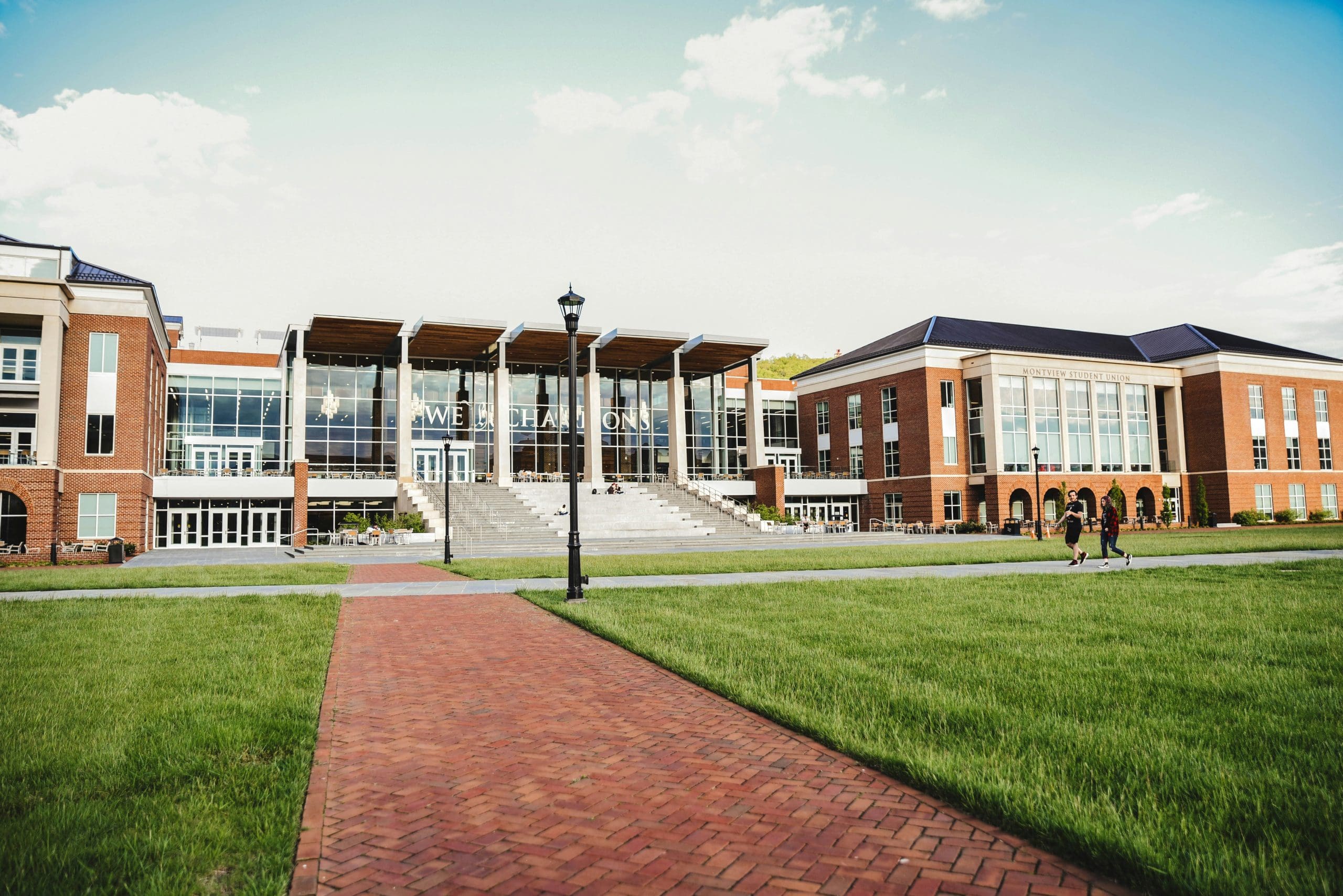 Campus university building with a glass-front entrance, wide stairs, and a green lawn; two students walk along a brick path in the foreground on a sunny day.